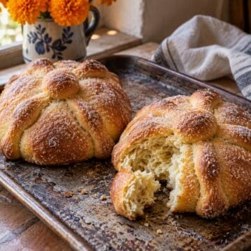 Two sugar coated pan de muerto loaves on a baking tray, one torn to show its soft crumb