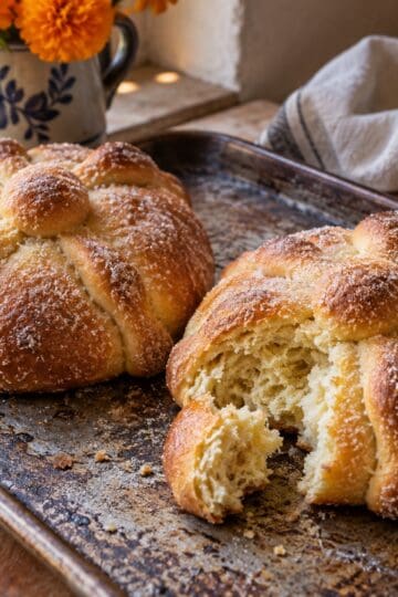 Two sugar coated pan de muerto loaves on a baking tray, one torn to show its soft crumb